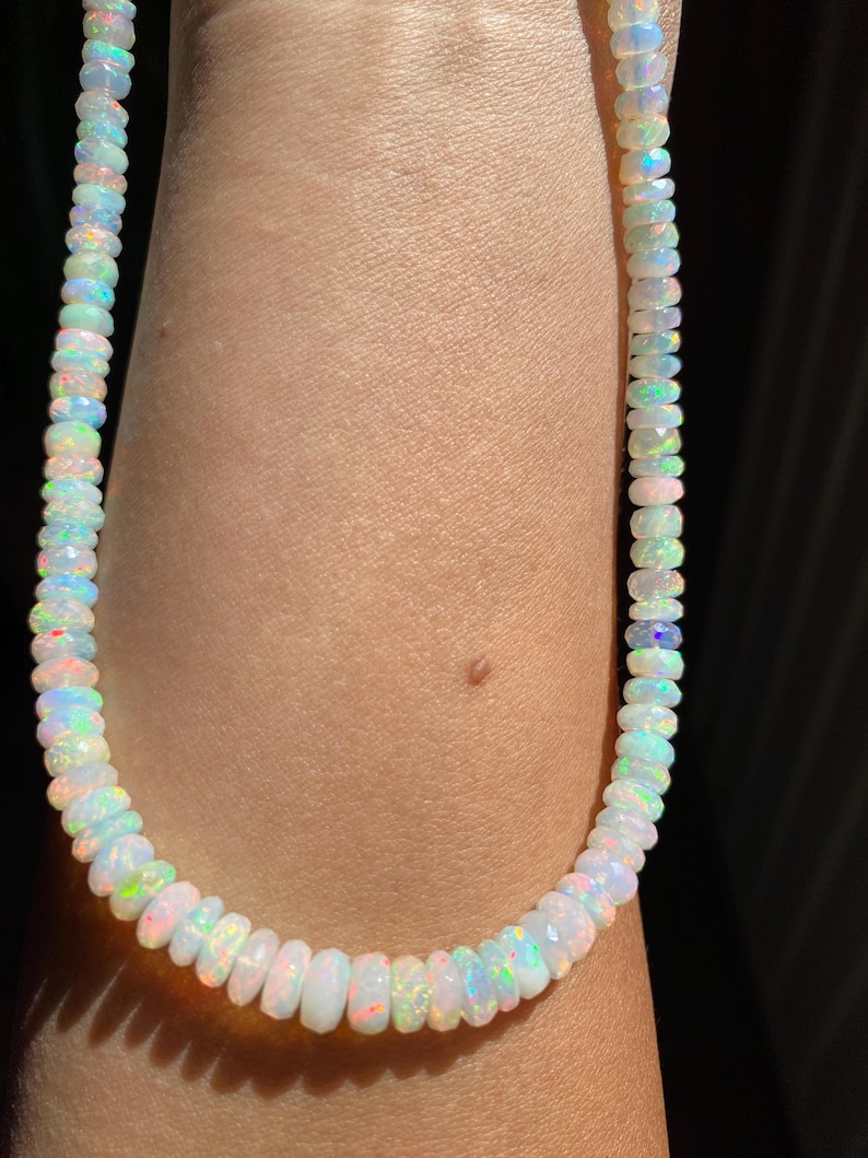 May include: A white opal bead necklace with iridescent flashes of colour. The beads are faceted and strung together, creating a delicate and eye-catching piece of jewellery. The necklace is shown on a person's arm.