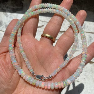 May include: A necklace made of small, faceted opal beads, with a silver clasp. The opals display a play of colours, including white, pink, and green. The necklace is shown against a hand.