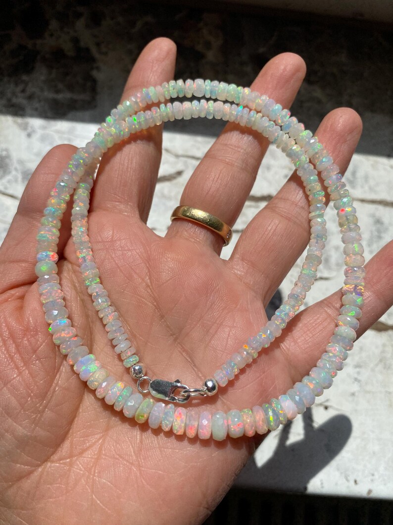 May include: A necklace made of small, faceted opal beads, with a silver clasp. The opals display a play of colours, including white, pink, and green. The necklace is shown against a hand.