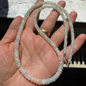 May include: A finely crafted opal necklace featuring a silver clasp. The necklace is made up of small, faceted opal beads, showcasing a spectrum of iridescent colours, including pink, green, and blue. The necklace is displayed on a hand.