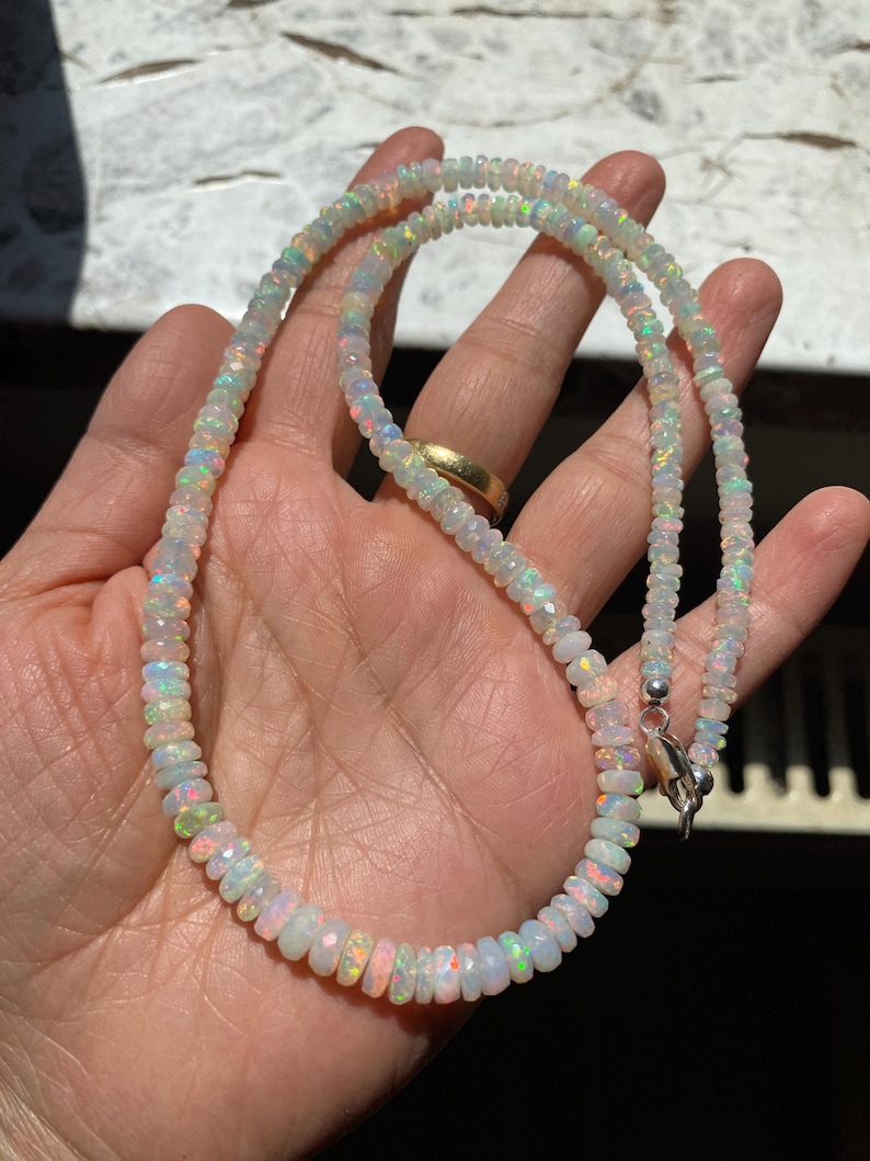 May include: A finely crafted opal necklace featuring a silver clasp. The necklace is made up of small, faceted opal beads, showcasing a spectrum of iridescent colours, including pink, green, and blue. The necklace is displayed on a hand.