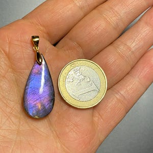 May include: A teardrop-shaped pendant with a gold bail, showcasing a vibrant purple and blue gemstone. The pendant is held next to a one Euro coin for size comparison. The gemstone has a polished surface with iridescent flashes.