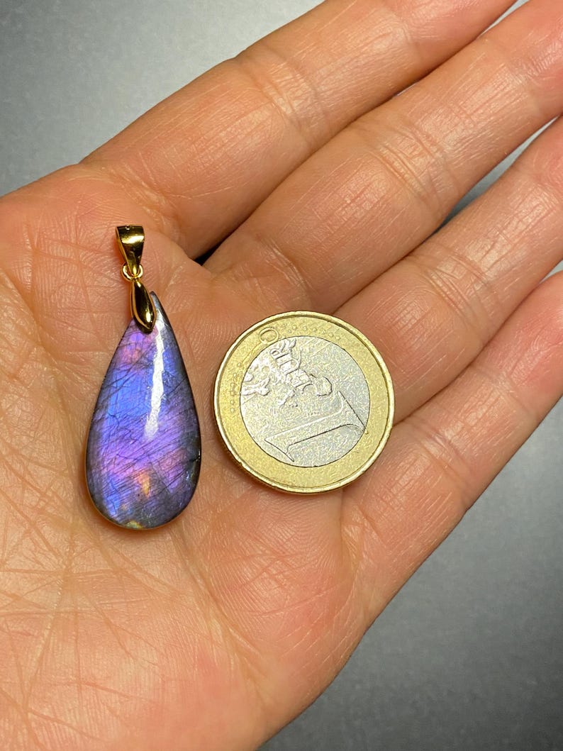 May include: A teardrop-shaped pendant with a gold bail, showcasing a vibrant purple and blue gemstone. The pendant is held next to a one Euro coin for size comparison. The gemstone has a polished surface with iridescent flashes.
