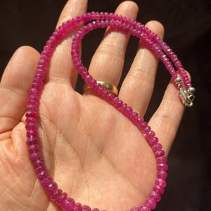Op de afbeelding: Een ketting bestaande uit kleine, gefacetteerde, diep roze-rode robijnkralen die aan elkaar zijn geregen. De streng is voorzien van een gepolijste, zilverkleurige metalen karabijnsluiting aan het uiteinde voor een veilige sluiting van dit sieraad.