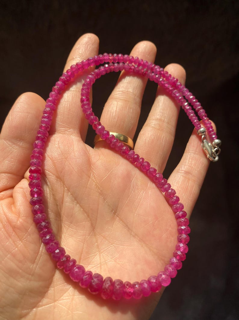 Op de afbeelding: Een ketting bestaande uit kleine, gefacetteerde, diep roze-rode robijnkralen die aan elkaar zijn geregen. De streng is voorzien van een gepolijste, zilverkleurige metalen karabijnsluiting aan het uiteinde voor een veilige sluiting van dit sieraad.