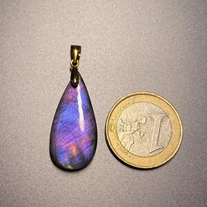 May include: A teardrop-shaped pendant with a gold-colored bail. The pendant is a vibrant mix of purple, blue, and orange hues. A one Euro coin is next to the pendant for size comparison. The pendant is likely a gemstone.