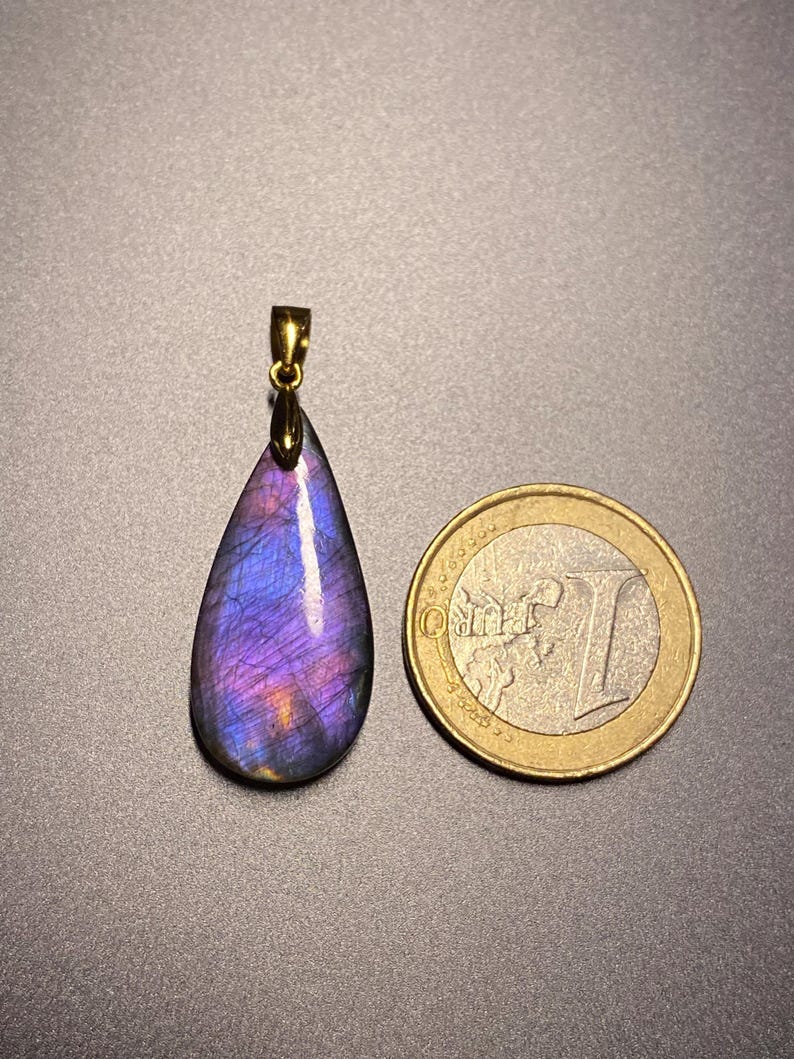 May include: A teardrop-shaped pendant with a gold-colored bail. The pendant is a vibrant mix of purple, blue, and orange hues. A one Euro coin is next to the pendant for size comparison. The pendant is likely a gemstone.