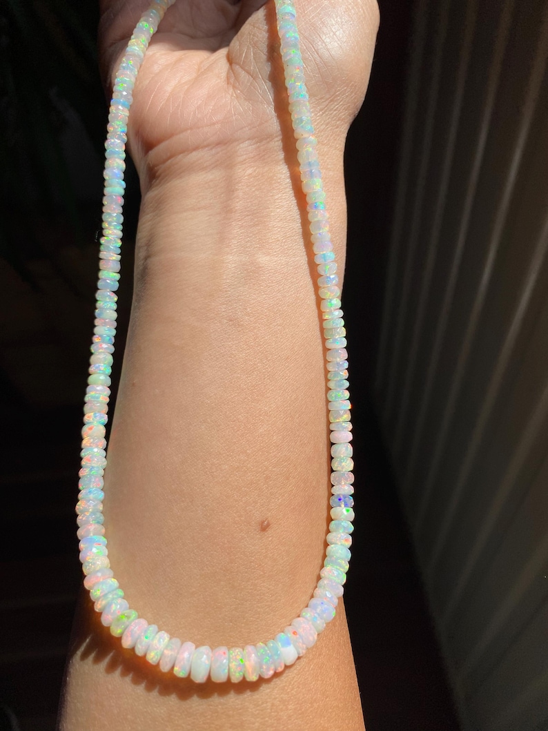 May include: A necklace made of small, iridescent opal beads. The beads show a range of colours, including white, blue, green, and pink, creating a subtle, shimmering effect. The necklace is strung together, forming a complete circle.