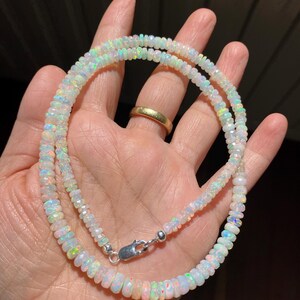 May include: A double-strand necklace made of iridescent opal beads. The beads are faceted and vary in size, connected with a silver-coloured clasp. The necklace is held in a hand, displaying its colourful play of light.