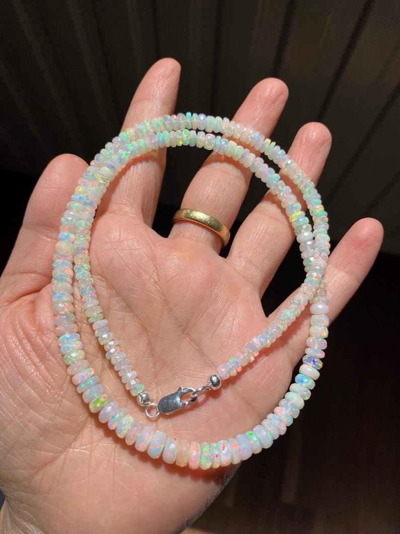 May include: A double-strand necklace made of iridescent opal beads. The beads are faceted and vary in size, connected with a silver-coloured clasp. The necklace is held in a hand, displaying its colourful play of light.