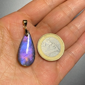 May include: A teardrop-shaped pendant with a gold bail, featuring a vibrant purple and blue gemstone. The pendant is displayed next to a Euro coin for size comparison. The gemstone has a polished surface and a gold setting.