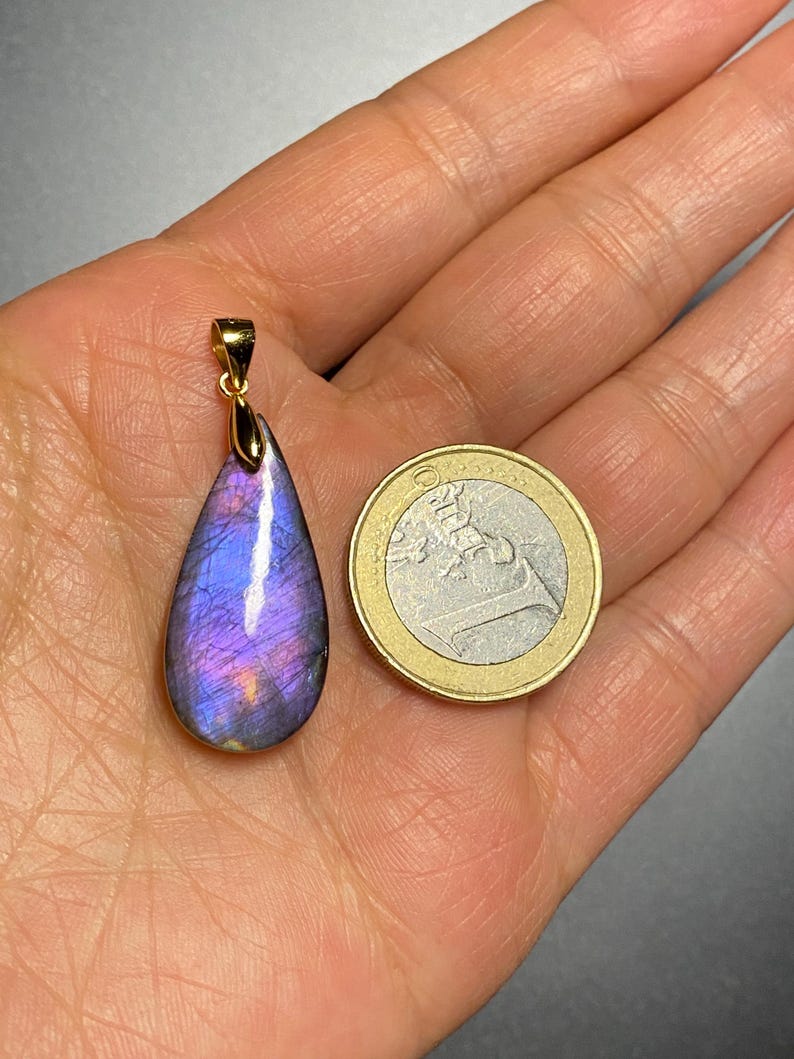 May include: A teardrop-shaped pendant with a gold bail, featuring a vibrant purple and blue gemstone. The pendant is displayed next to a Euro coin for size comparison. The gemstone has a polished surface and a gold setting.