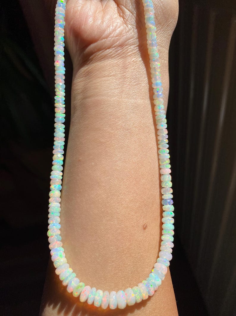 May include: A necklace made of small, iridescent opal beads. The beads show a range of colours, including white, blue, green, and pink. The necklace is displayed against a skin tone background.