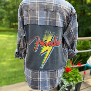 May include: A long-sleeve button-down flannel shirt in a blue, grey, and white plaid pattern. The back features a large, rectangular black fabric patch with a graphic of a yellow lightning bolt and the red Fender logo printed over it.