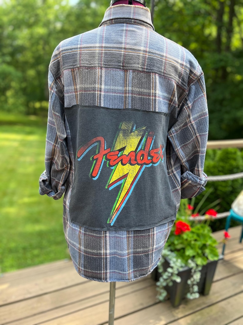 May include: A long-sleeve button-down flannel shirt in a blue, grey, and white plaid pattern. The back features a large, rectangular black fabric patch with a graphic of a yellow lightning bolt and the red Fender logo printed over it.