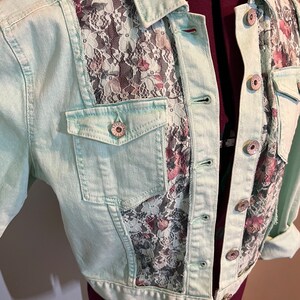 May include: A light green denim jacket with a floral lace overlay on the front and back. The jacket has a button closure and two pockets.