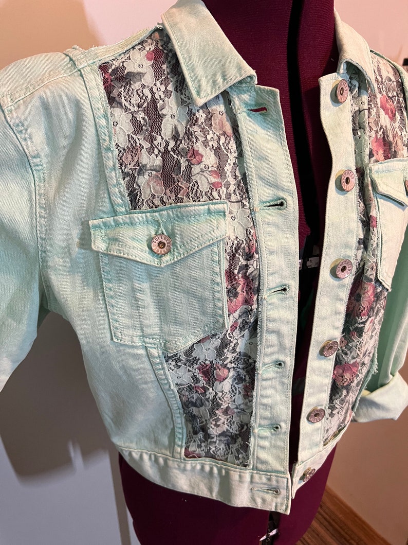 May include: A light green denim jacket with a floral lace overlay on the front and back. The jacket has a button closure and two pockets.