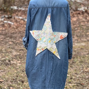 May include: A blue denim shirt with a large patchwork star on the back. The star is made of various fabrics in shades of white, pink, yellow, and green.