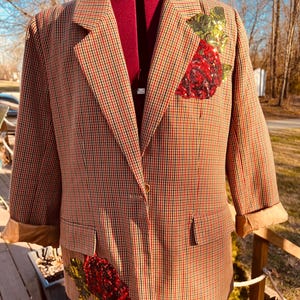May include: Brown, red, and black plaid blazer with a single gold button closure. The blazer features a sequined red rose design on the chest and lower left pocket. The sleeves are cuffed, revealing a tan lining. The blazer is displayed on a mannequin.
