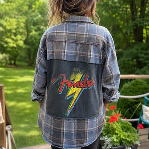 May include: A long-sleeve button-down flannel shirt in a blue and grey plaid pattern. The back features a large, rectangular black fabric patch with a graphic of a yellow lightning bolt and the red Fender logo with blue and green outlines.