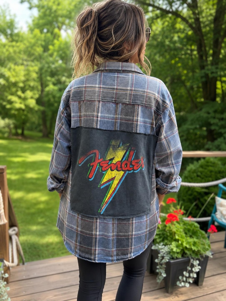May include: A long-sleeve button-down flannel shirt in a blue and grey plaid pattern. The back features a large, rectangular black fabric patch with a graphic of a yellow lightning bolt and the red Fender logo with blue and green outlines.