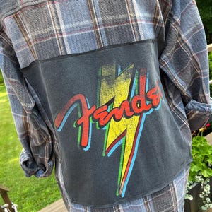 May include: A blue and grey plaid long-sleeve button-down shirt featuring a large rectangular black fabric patch sewn onto the back. The patch displays a distressed yellow lightning bolt graphic behind the red Fender brand logo text.