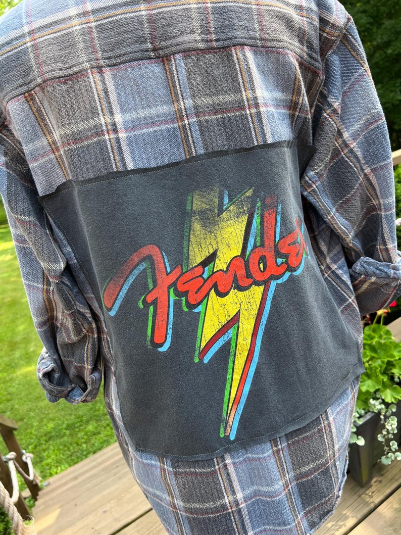 May include: A blue and grey plaid long-sleeve button-down shirt featuring a large rectangular black fabric patch sewn onto the back. The patch displays a distressed yellow lightning bolt graphic behind the red Fender brand logo text.