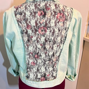 May include: A light blue denim jacket with a floral lace back panel. The lace is white with pink and grey flowers.