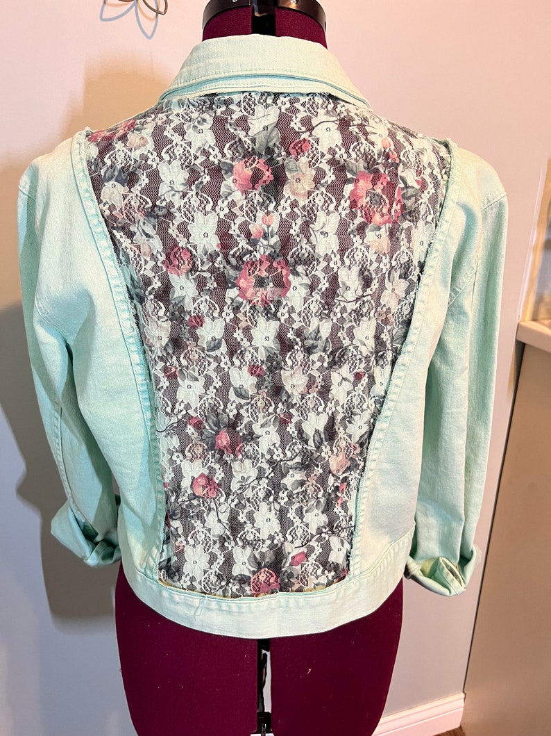 May include: A light blue denim jacket with a floral lace back panel. The lace is white with pink and grey flowers.