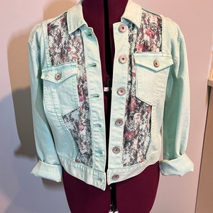 May include: A light blue denim jacket with a floral lace panel on the back. The jacket has a button closure and two front pockets.