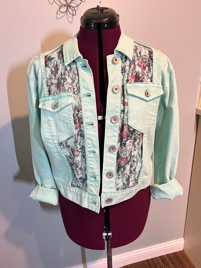 May include: A light blue denim jacket with a floral lace panel on the back. The jacket has a button closure and two front pockets.