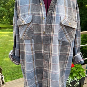 May include: A long-sleeved button-down shirt featuring a plaid pattern in shades of blue, grey, and white with thin red and yellow accent lines. The shirt includes a standard collar, two chest pockets with buttoned flaps, and cuffed sleeves.