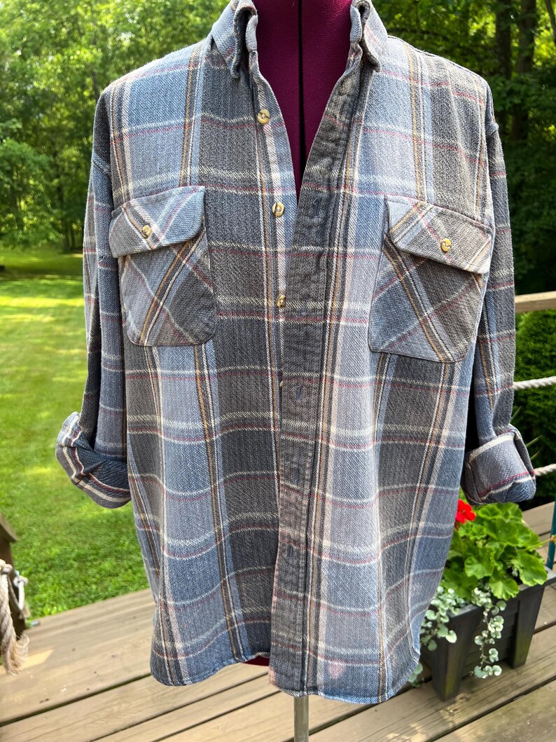 May include: A long-sleeved button-down shirt featuring a plaid pattern in shades of blue, grey, and white with thin red and yellow accent lines. The shirt includes a standard collar, two chest pockets with buttoned flaps, and cuffed sleeves.