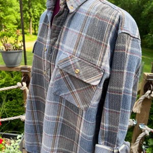May include: A long-sleeved button-down shirt featuring a plaid pattern in shades of blue, grey, and white with thin red accent lines. The shirt includes a pointed collar, a single chest pocket with a button closure, and cuffs that are rolled up.