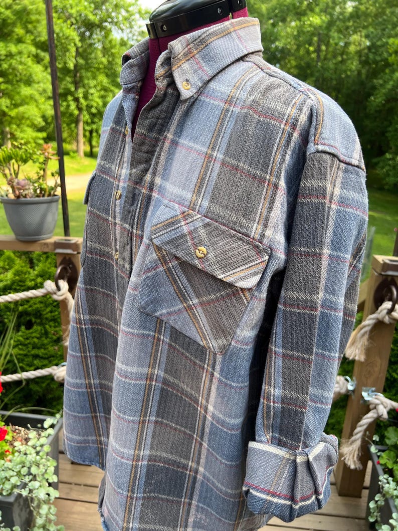 May include: A long-sleeved button-down shirt featuring a plaid pattern in shades of blue, grey, and white with thin red accent lines. The shirt includes a pointed collar, a single chest pocket with a button closure, and cuffs that are rolled up.