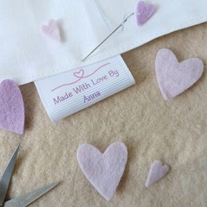May include: A white fabric label with the purple text Made With Love By Anna and a heart icon, attached to white fabric with a silver sewing needle. Several light purple felt hearts are scattered around, and silver metal scissors are visible.