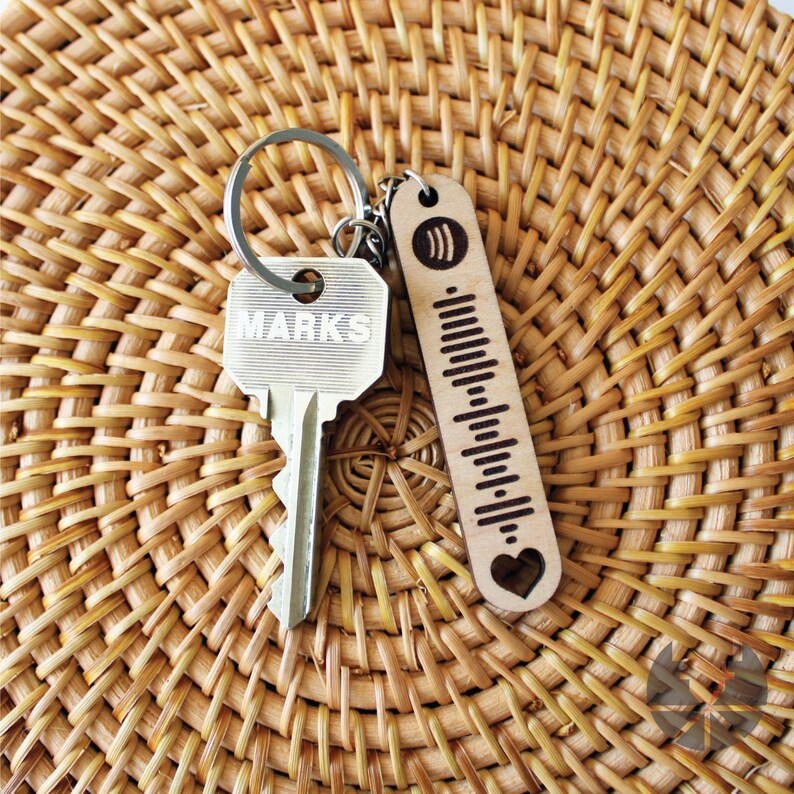 May include: A silver key with the word "MARKS" engraved on it is attached to a wooden keychain with a heart and a musical note design. The keychain is engraved with a series of vertical lines.