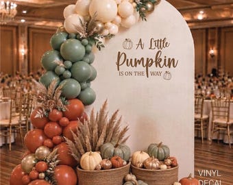 May include: A beige arched backdrop featuring the text A Little Pumpkin Is On The Way, decorated with a balloon garland in cream, sage green, and terracotta, accented with pampas grass and assorted pumpkins in woven baskets.