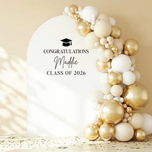 A detailed interior party decor mockup featuring a cream-beige arched panel against a white textured wall with distinctive window shadows. The panel displays customizable text: "CONGRATULATIONS", the script name, a graduation cap icon, and "CLASS OF 2026". An asymmetrical organic balloon garland with cream, pearl white, and gold-marbled balloons cascades down the right side of the arch. The rustic wooden plank floor is scattered with gold star and dot confetti.