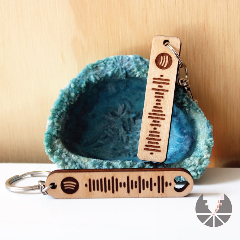 May include: Two wooden keychains with a Spotify logo and a barcode design. The keychains are on a blue and white background.