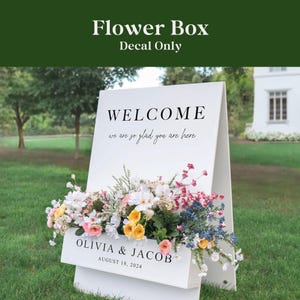 May include: A white flower box sign with the text "WELCOME we are so glad you are here". The sign also includes the names "OLIVIA & JACOB" and the date "AUGUST 18, 2024". The flower box is filled with colorful flowers.