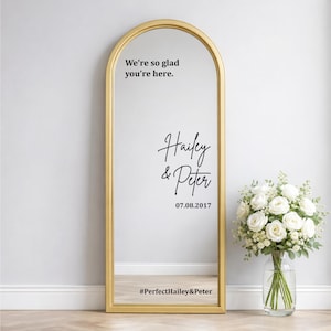 Custom wedding mirror decal mockup on a silver arched floor mirror. Black text reads Hello Darling, You look incredible now go grab a drink, and Andrew plus Mina January 12 2024. Next to a white vase with pampas grass on a wood floor.