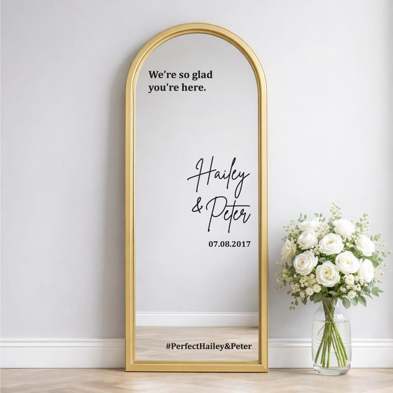 Custom wedding mirror decal mockup on a silver arched floor mirror. Black text reads Hello Darling, You look incredible now go grab a drink, and Andrew plus Mina January 12 2024. Next to a white vase with pampas grass on a wood floor.
