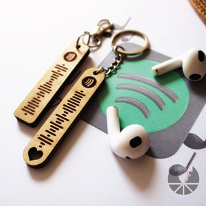May include: Two wooden keychains with a heart and music note design. The keychains are engraved with a Spotify barcode. The keychains are attached to a silver key ring. The keychains are on a white surface with a green Spotify logo and white earbuds.
