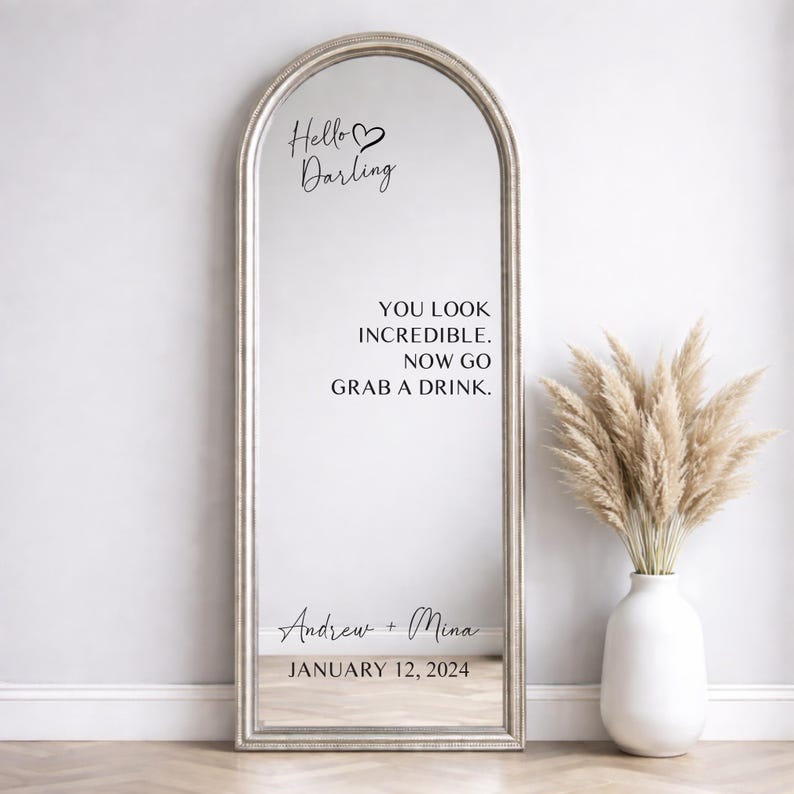 Custom wedding mirror decal mockup on a silver arched floor mirror. Black text reads Hello Darling, You look incredible now go grab a drink, and Andrew plus Mina January 12 2024. Next to a white vase with pampas grass on a wood floor.