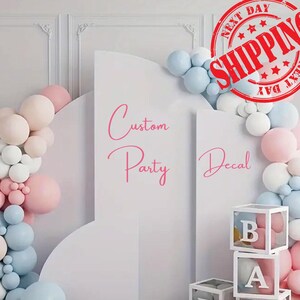 May include: A white arched backdrop display featuring the pink text Custom Party Decal, decorated with a cluster of light blue, white, and pale pink balloons, and stacked white transparent boxes containing balloons with letters B and A.