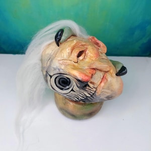 May include: A handcrafted sculpture of a fantastical creature's head. The sculpture features a detailed face with a large eye, white hair, and green and black accents. The head rests on a rounded base, with a color palette of beige, red, and green.