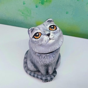 May include: A gray and white ceramic cat figurine with orange eyes. The cat is sitting with its tail curled around its body.