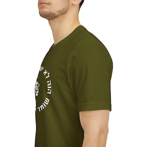 Op de afbeelding: Een olijfgroen T-shirt met korte mouwen en een cirkelvormige witte afbeelding op de borst. Het ontwerp bevat een gestileerd leeuwengezicht in het midden, omringd door Hebreeuwse tekst in een cirkel, gedrukt in een vet, strak wit lettertype.