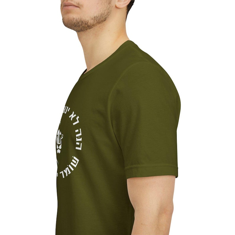 Op de afbeelding: Een olijfgroen T-shirt met korte mouwen en een cirkelvormige witte afbeelding op de borst. Het ontwerp bevat een gestileerd leeuwengezicht in het midden, omringd door Hebreeuwse tekst in een cirkel, gedrukt in een vet, strak wit lettertype.